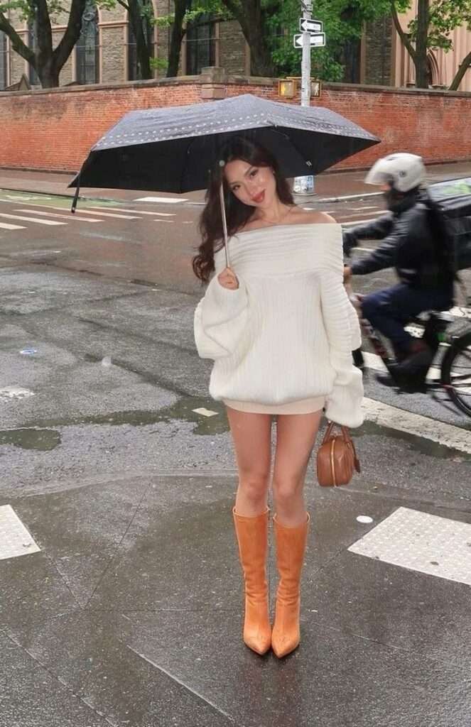 This look centers on a dramatic, oversized off-the-shoulder white knit sweater, elegantly worn as a playful micro-mini dress that emphasizes the bare leg silhouette. The cozy volume is sharply contrasted by a striking pair of pointed-toe knee-high boots in a rich, warm camel hue, anchoring the outfit with a directional color pop. Paired with a structured brown handbag and a black umbrella, this ensemble masters effortless city chic for transitional, wet weather.