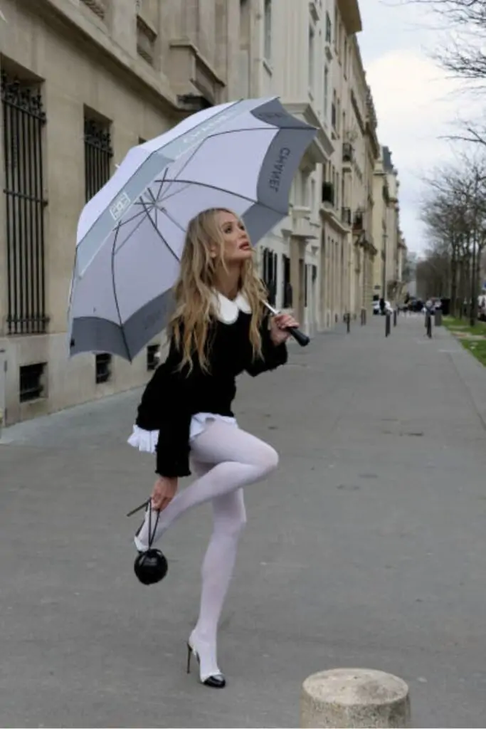 This look gives a playful twist to classic silhouettes, featuring a black micro-mini dress or sweater with a sharp white Peter Pan collar and a voluminous white ruffled underlayer, emphasizing the hemline. The ensemble is defined by the striking contrast of opaque white tights paired with chic white cap-toe slingback heels, creating a strong visual line, and is accessorized with a branded umbrella and a spherical novelty bag.