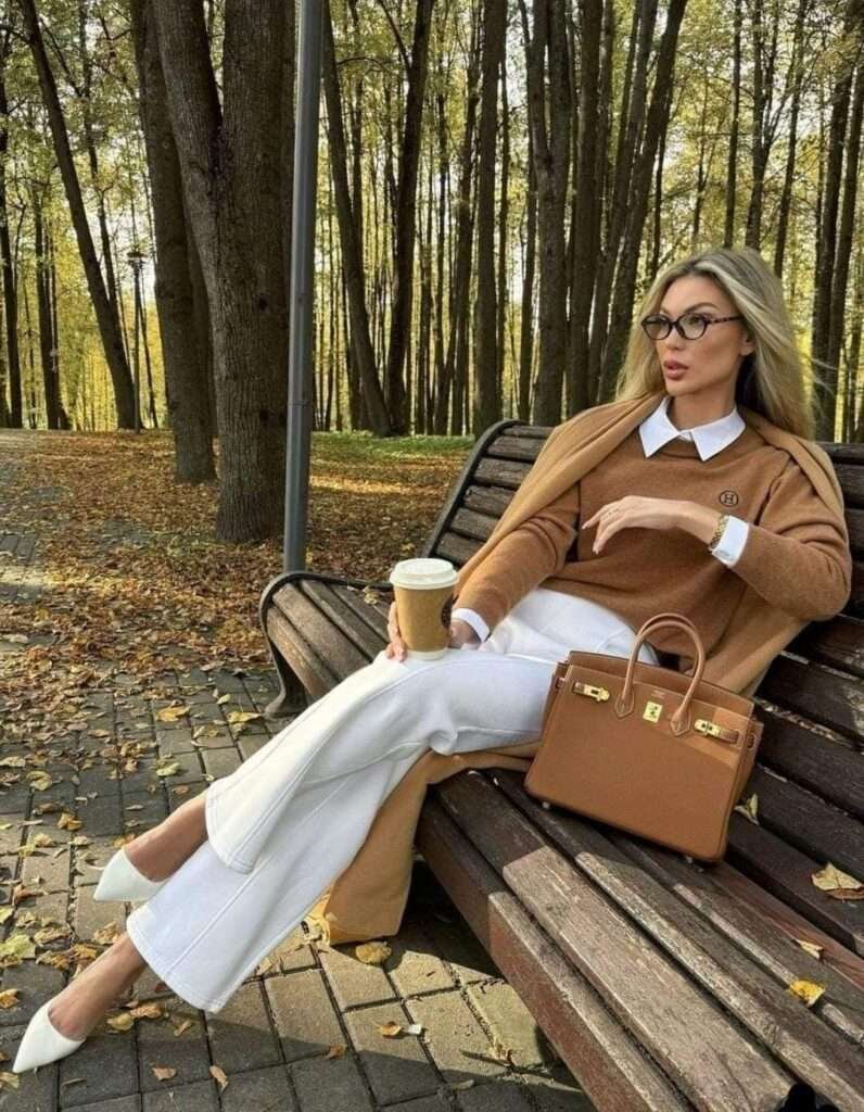 This polished ensemble perfectly balances seasonal tones with crisp contrasts, featuring a rich camel-toned knit sweater layered elegantly over a white collared shirt, and paired with chic high-waisted white wide-leg trousers. The look is completed with sharp white pointed-toe pumps and a structured Hermès Birkin-style bag in matching cognac leather, embodying quiet luxury suitable for a fall park setting.