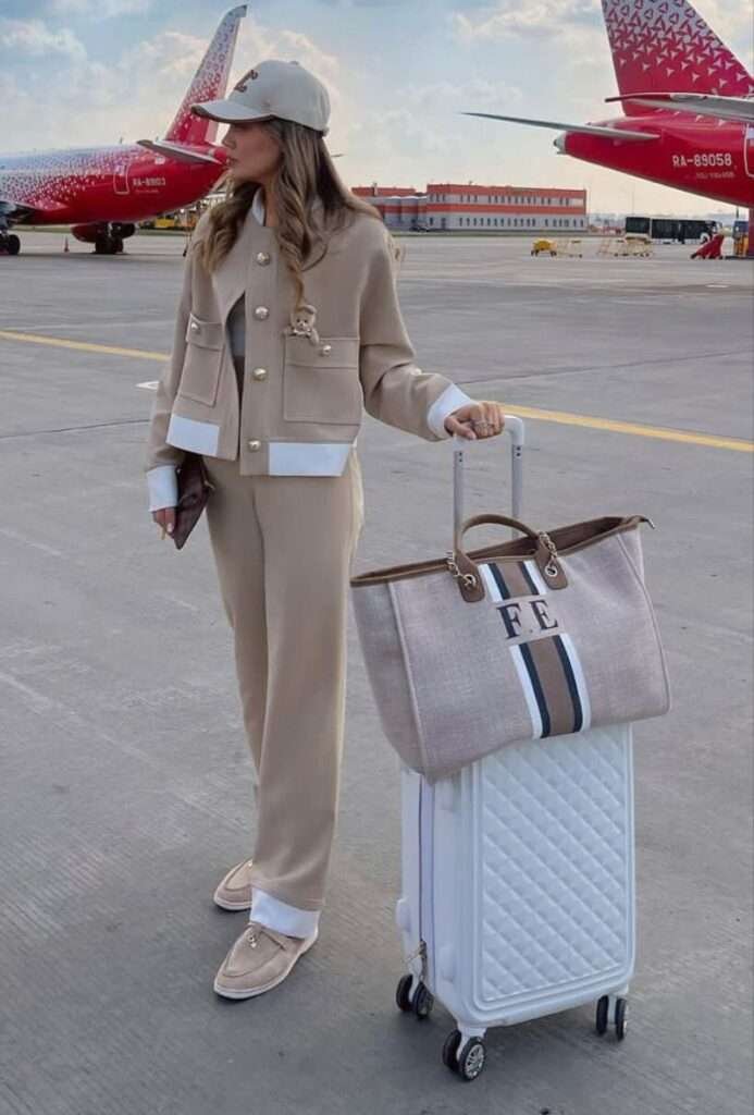 This polished ensemble perfectly balances seasonal tones with crisp contrasts, featuring a rich camel-toned knit sweater layered elegantly over a white collared shirt, and paired with chic high-waisted white wide-leg trousers. The look is completed with sharp white pointed-toe pumps and a structured Hermès Birkin-style bag in matching cognac leather, embodying quiet luxury suitable for a fall park setting.