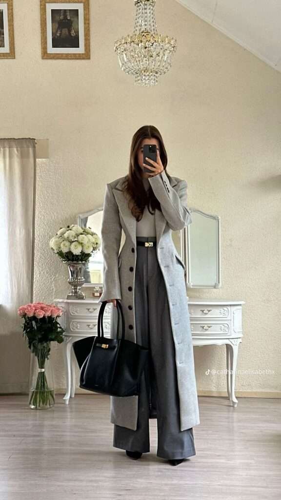 The look centers on sharp, elongated lines and tonal gray dressing, featuring an impactful light gray maxi coat with defined shoulder pads and a pronounced collar, cinched at the waist. The coat is layered over dark gray pleated wide-leg trousers and a subtly-colored top, with a striking gold-buckle belt serving as the focal point to create a strong, hourglass silhouette. The ensemble is impeccably finished with a classic, structured black leather tote bag, embodying timeless power dressing.