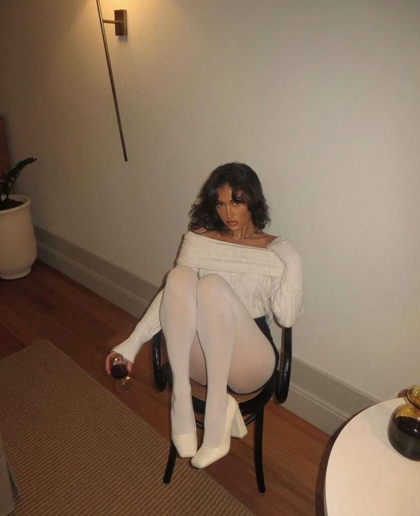 This sophisticated style centers on soft textures and striking monochromatic legwear, featuring a creamy off-the-shoulder ribbed knit sweater that elegantly drapes across the collarbones. The look is anchored by high-coverage opaque white tights paired with matching chunky-heeled pumps, creating an unbroken, statuesque line that contrasts sharply with the subtle dark element of the mini-skirt beneath, offering a perfect blend of comfort and quiet luxury for an intimate evening.