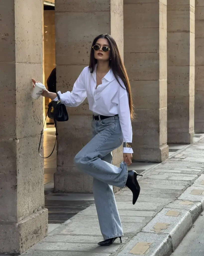 Stylish woman with long straight dark hair and round gold-framed sunglasses posing dynamically against a classic stone column in an upscale urban setting, wearing an oversized white button-down shirt with deep V-neck unbuttoned at the top, high-waisted light wash wide-leg jeans cinched with a slim black belt, black pointed-toe kitten heels, a small black structured handbag, and holding a takeaway coffee cup, exuding confident and chic casual spring fashion for 2026.