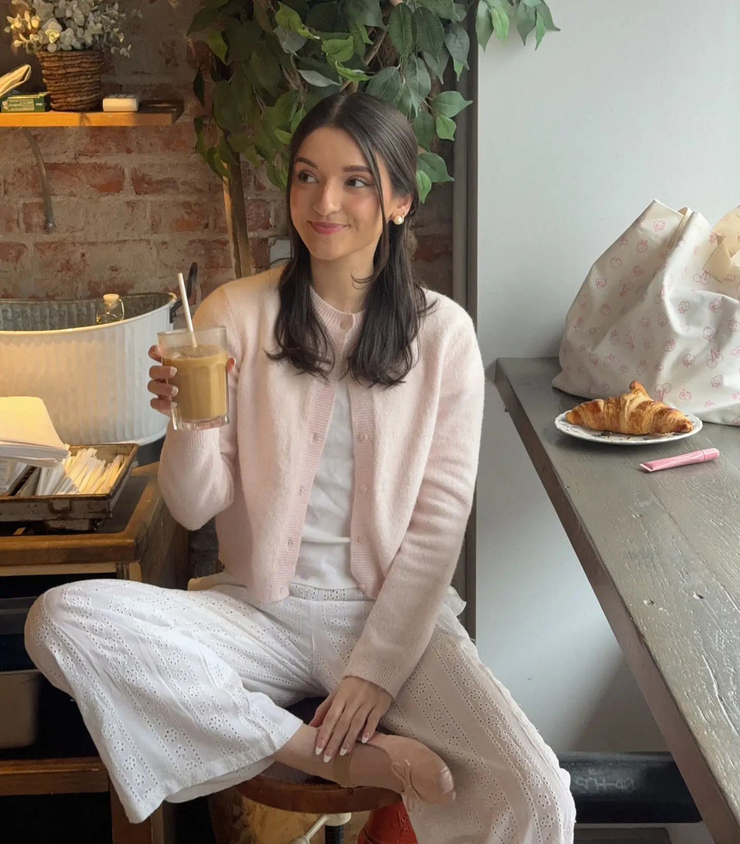 Obsessed with this dreamy brunch-ready spring look for 2026: a cloud-like pastel pink cardigan layered over crisp white separates with intricate eyelet detailing creates the coziest yet most polished vibe, finished with dainty ballet flats, a sweet flower in the hair, and a tall iced latte. Captured in a light-filled café surrounded by cascading greenery and fresh blooms, the outfit perfectly captures that soft, romantic spring aesthetic – think leisurely mornings, coffee dates, or effortless weekend elegance.