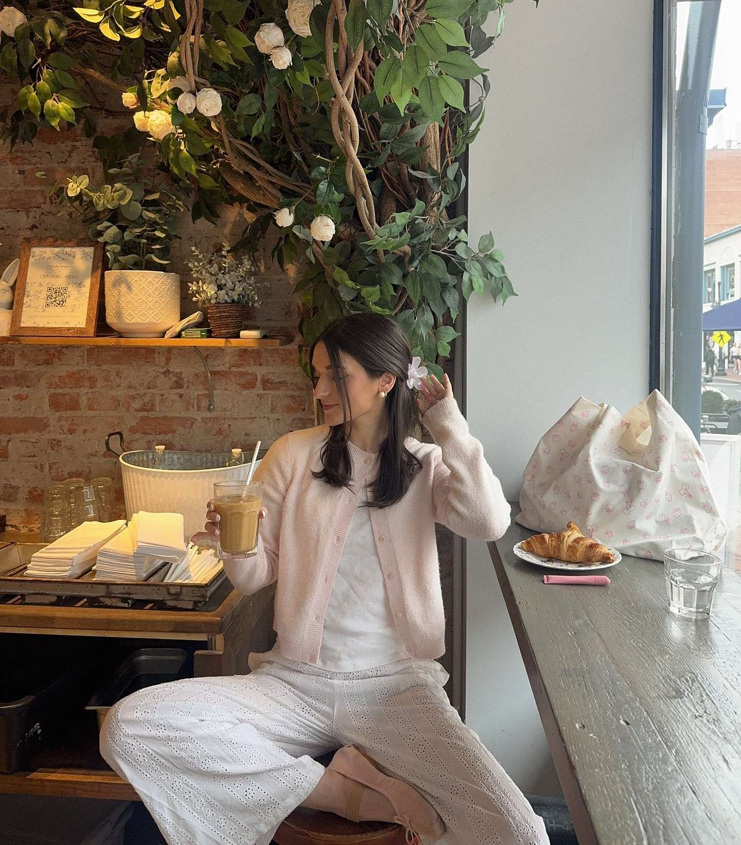 Woman with long dark wavy hair adorned with a delicate white flower clip, sitting relaxed on a wooden stool at a charming café table, wearing a soft pastel pink mohair cardigan over a simple white top, paired with matching high-waisted white eyelet lace wide-leg trousers, nude ballet flats, holding a tall iced latte in a glass, with a croissant on a plate, a large floral-patterned canvas tote bag, and lush green foliage wall with white roses behind her, evoking a serene, feminine, and dreamy spring aesthetic for 2026.