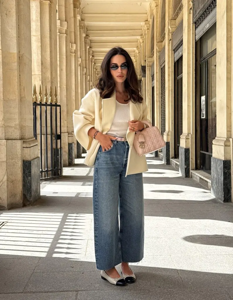Confident woman with long wavy dark hair and dark rectangular sunglasses standing in a grand arched colonnade with classic stone columns and marble details, wearing an oversized cream blazer with wide lapels over a fitted white ribbed tank top, high-waisted light wash wide-leg jeans with frayed hems, a thin pink belt, two-tone cream and black pointed-toe ballet flats, and holding a small pink quilted leather handbag with gold hardware, exuding a timeless, sophisticated, and effortlessly chic neutral spring aesthetic for 2026. This polished Parisian-inspired spring ensemble is 2026 wardrobe essential: a slouchy cream blazer with dramatic sleeves drapes perfectly over a simple white ribbed tank, paired with high-waisted wide-leg light denim jeans cinched by a delicate pink belt and grounded by chic two-tone ballet flats.