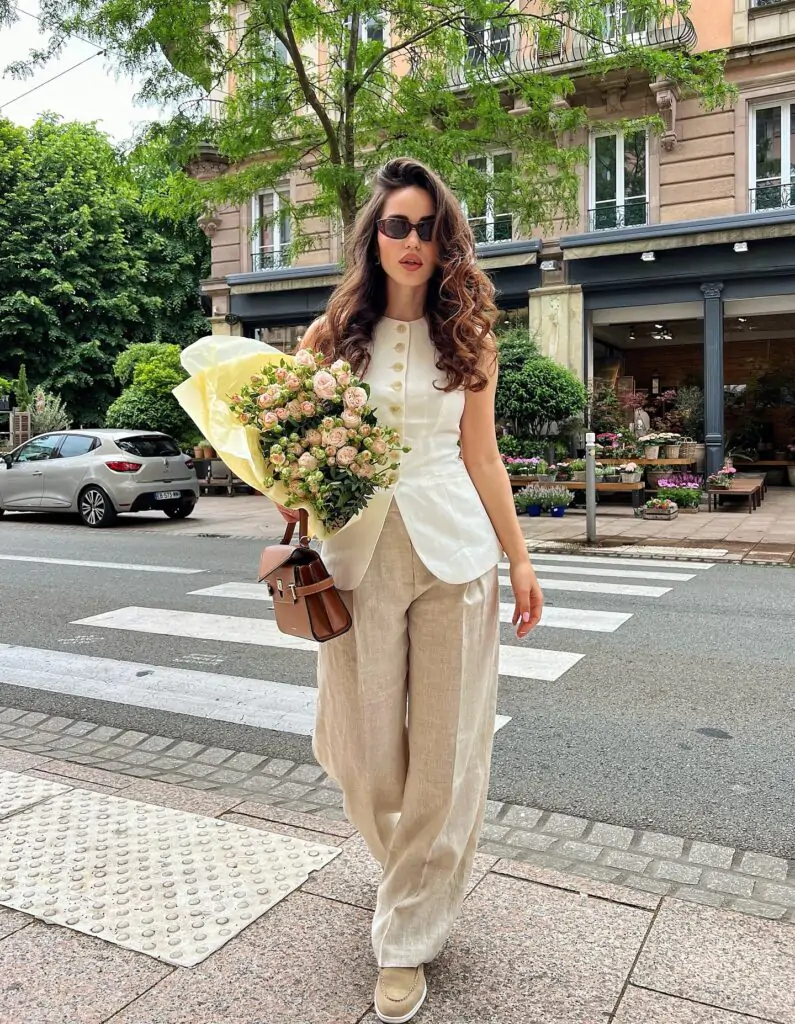 This clean and luxurious neutral spring ensemble is made for 2026: a tailored sleeveless cream vest with subtle gold buttons flows perfectly into high-waisted wide-leg linen trousers, grounded by simple nude sneakers and finished with a structured tan bag plus a romantic bouquet of soft pink roses. Captured crossing a charming street lined with blooming flower stalls and elegant buildings, the outfit radiates timeless sophistication and pure spring aesthetic – perfect for market mornings, casual lunches, or effortless weekend elegance.