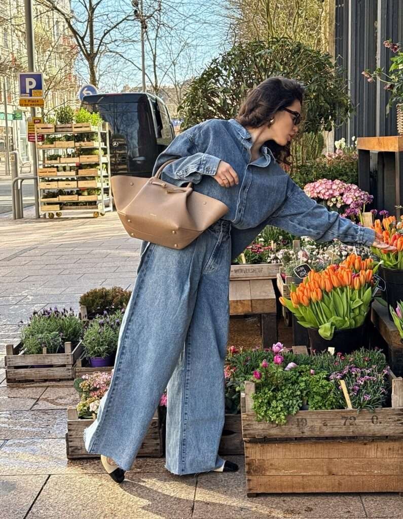 Woman in oversized blue denim shirt, wide-leg jeans, beige studded handbag, and sunglasses, selecting orange tulips at an outdoor flower market, capturing a relaxed casual spring fashion vibe for 2026.