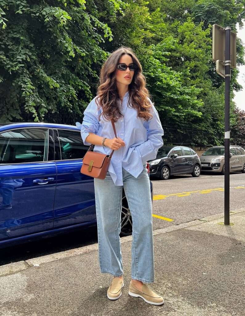 Blonde woman with hair in a high bun decorated with a yellow flower, walking gracefully away from the camera under a tunnel of blooming pink cherry blossom trees in an urban park, wearing an off-shoulder white open-back lace knit sweater, high-waisted light blue wide-leg jeans cuffed at the ankles, white pointed-toe ballet flats, and carrying a large woven straw tote bag with brown leather straps over her shoulder plus a bouquet of bright yellow sunflowers wrapped in kraft paper, embodying a lighthearted and feminine spring aesthetic for 2026.