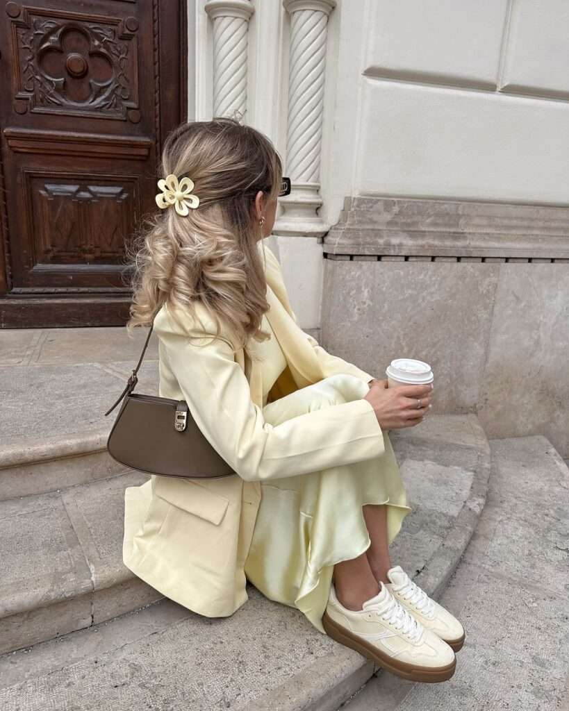 Side view of a woman with long wavy blonde hair adorned with a cream flower hair clip, wearing a chic light yellow oversized blazer and matching asymmetrical midi skirt, paired with cream low-top sneakers, holding a to-go coffee cup and a small brown shoulder bag, sitting casually on stone steps in front of an ornate wooden door with carved details and white spiral columns, radiating a soft and elegant spring aesthetic for 2026.
