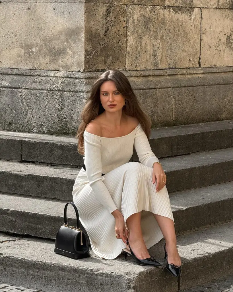 Elegant woman with long wavy brown hair and subtle makeup sitting gracefully on wide stone steps of a historic building, wearing an off-the-shoulder cream ribbed knit midi dress with long sleeves and pleated details, cinched with a slim black belt, black pointed-toe slingback heels, and a small black structured handbag placed beside her, exuding a timeless and feminine classy spring look for 2026.
