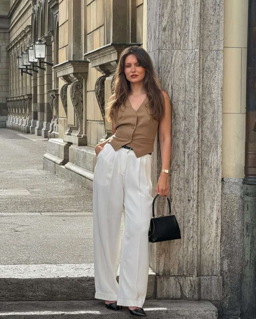 This clean-lined, luxurious spring moment is pure 2026 inspiration: a sleeveless taupe vest with delicate button details layers beautifully over a simple base and flows into high-waisted wide-leg ivory trousers that drape with effortless grace. Paired with glossy black pumps, a chic black handbag, and subtle gold jewelry, the outfit feels polished and modern yet completely timeless. Captured leaning against classic stone columns in a stunning colonnade, it perfectly captures that soft, elegant spring aesthetic – ideal for city days, gallery visits, or elevated everyday style.