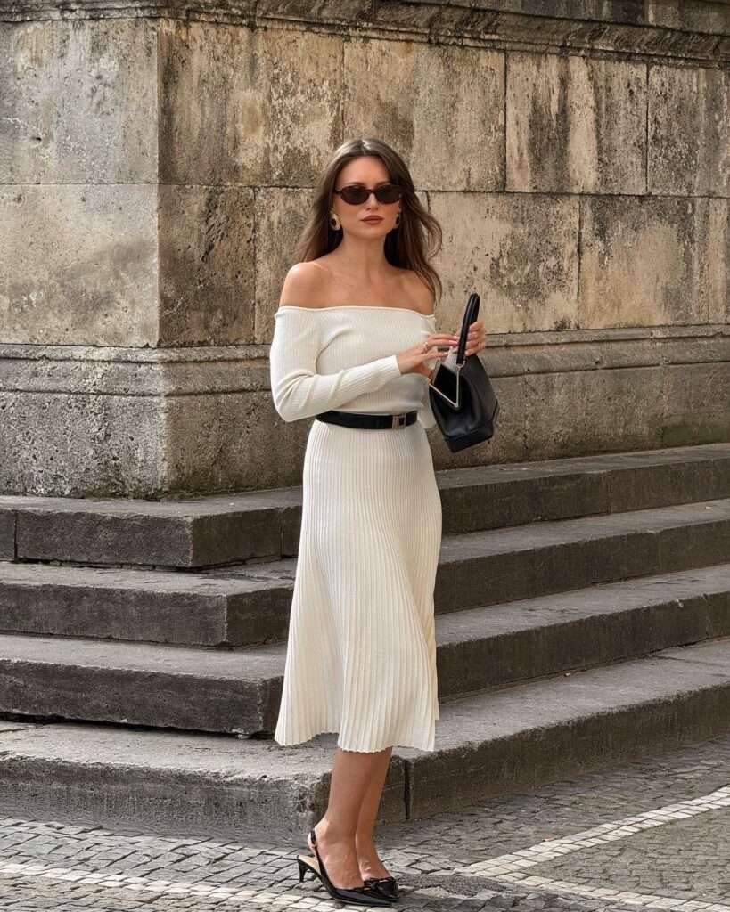 This stunning neutral spring outfit screams 2026 sophistication: a figure-skimming off-shoulder ribbed cream midi dress with long sleeves, accented by a thin black belt, glossy black slingbacks, and a chic black handbag. Leaning against ancient stone architecture on cobblestone streets, the look captures pure spring aesthetic with its clean lines and luxurious texture – ideal for dates, gallery visits, or elevated everyday wear. Save this classy spring vibe to recreate the romance of the season!