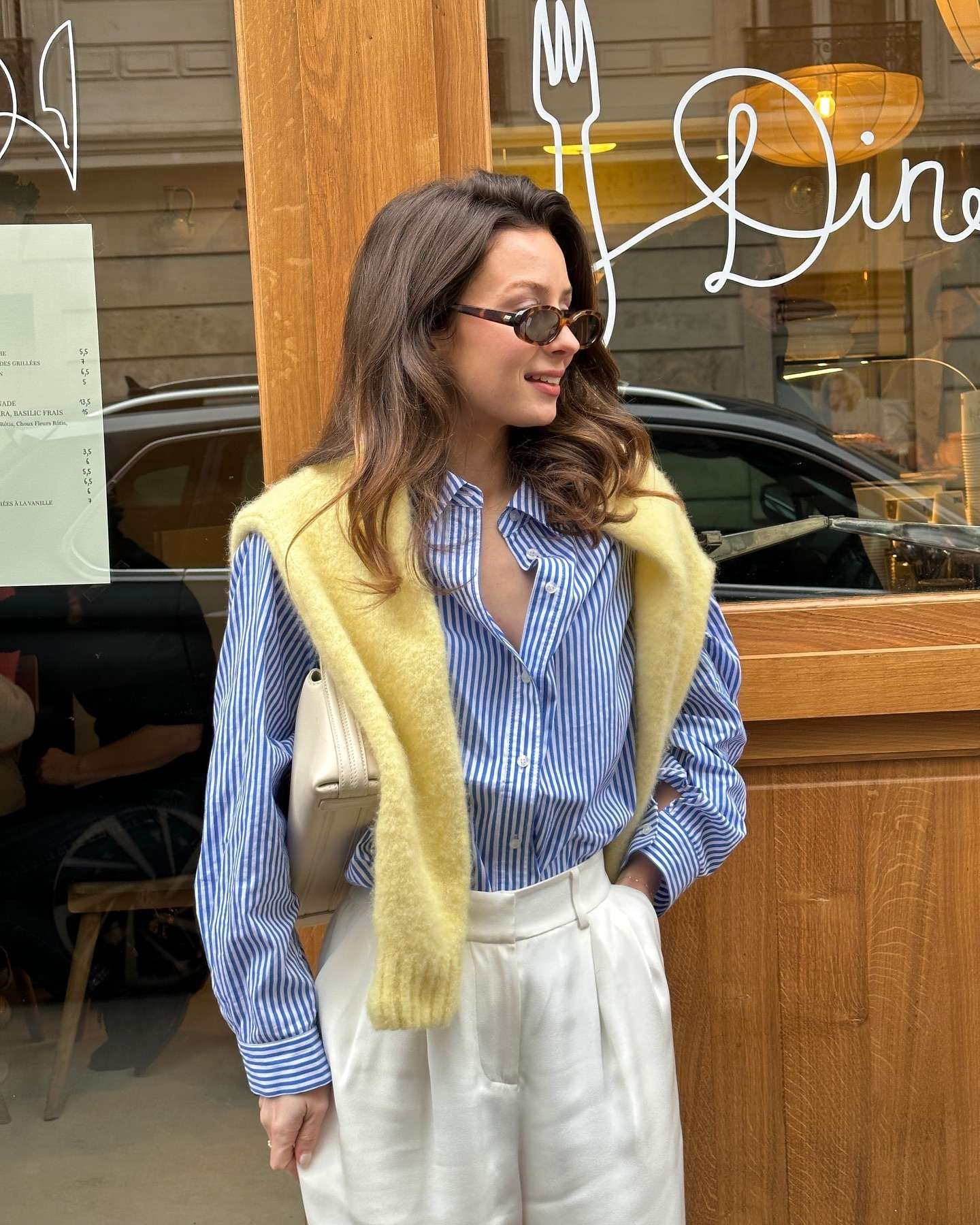 This soft and dreamy spring ensemble is 2026 wardrobe goals: a fluffy light yellow mohair vest adds the coziest layer over a crisp blue-and-white striped button-down, paired beautifully with high-waisted cream pleated trousers and a sleek beige bag. Captured smiling outside a cute Parisian café with warm lighting and handwritten menus, the outfit radiates pure spring aesthetic – light, feminine, and effortlessly elegant. Perfect for brunch dates, casual outings, or adding a touch of luxury to everyday style.