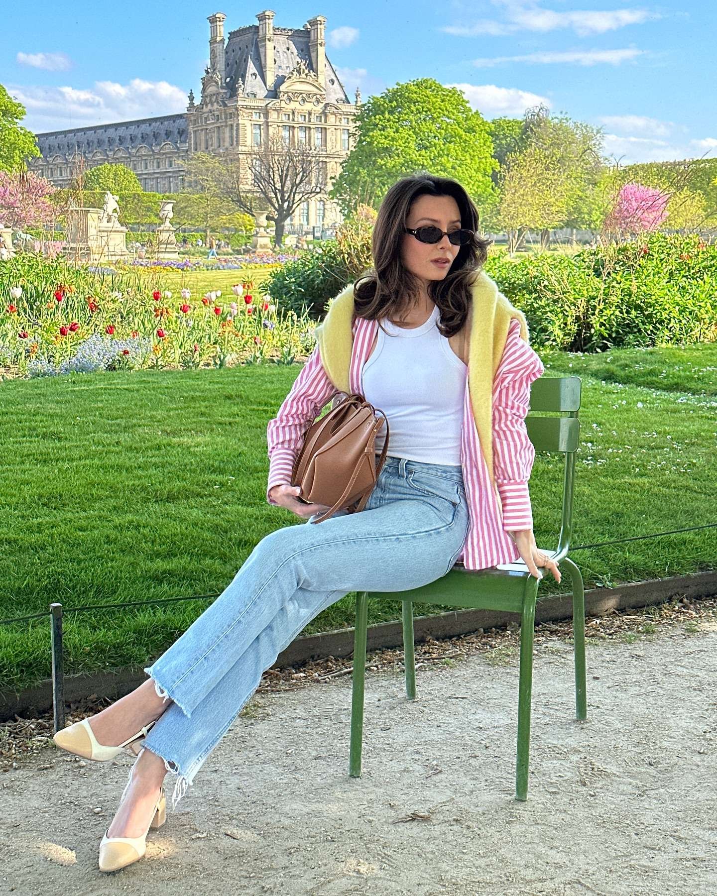 Elegant woman with long wavy dark hair and sleek black oval sunglasses sitting relaxed on a classic green metal park bench in the Tuileries Garden, wearing a light pink-and-white striped button-down shirt layered under a fluffy pastel yellow mohair vest, a white tank top underneath, high-waisted light wash wide-leg jeans with frayed hems, two-tone beige and cream pointed-toe block-heel pumps, and holding a structured tan leather backpack-style bag, with the iconic Louvre Pyramid and blooming spring tulips in vibrant flower beds behind her, embodying a soft, luxurious, and timeless spring aesthetic for 2026.