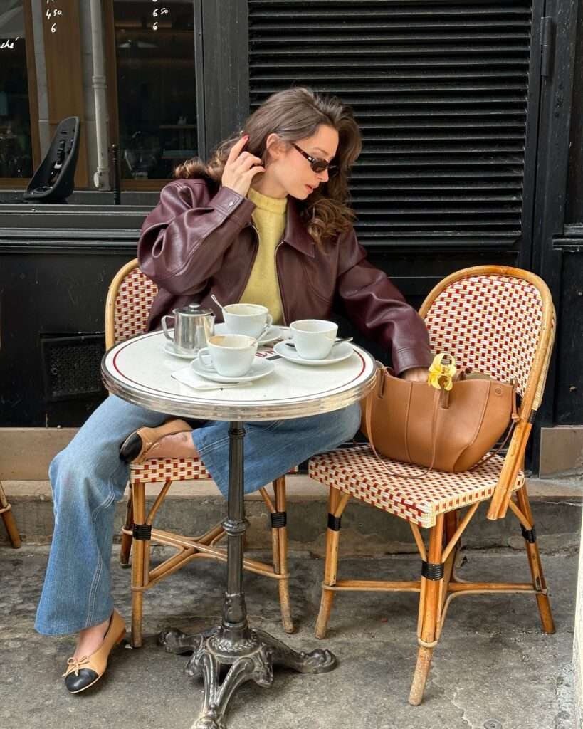 Sophisticated woman with long wavy brown hair and slim black sunglasses sitting at a classic Parisian café table with black shutters, wearing a rich burgundy leather jacket over a soft mustard yellow turtleneck sweater, high-waisted light wash wide-leg jeans cuffed at the ankles, two-tone beige and black pointed-toe ballet flats, and a structured tan leather tote bag with a yellow flower accent placed on the wicker chair beside her, enjoying coffee service on a small round marble-top table, capturing an effortlessly cool and luxurious casual spring fashion vibe for 2026.