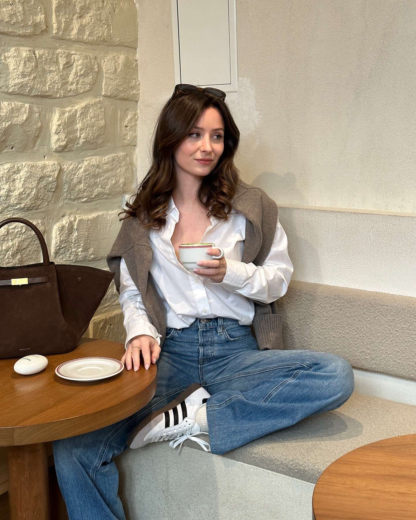 Woman with long wavy brown hair and sunglasses perched on her head sitting casually on a cozy café bench with exposed brick walls, wearing an oversized gray wool vest layered over a crisp white button-down shirt unbuttoned at the top, high-waisted light wash wide-leg jeans, white Adidas sneakers with black stripes, white crew socks, holding a colorful rimmed coffee cup, with a large brown leather tote bag and a small white Chanel soap bar on the wooden table beside her, radiating a relaxed, minimalist, and effortlessly chic casual spring fashion vibe for 2026.