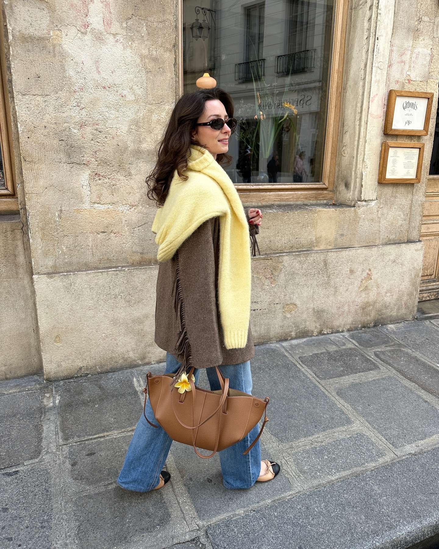 Stylish woman with long wavy dark hair and black oval sunglasses walking along a charming Parisian street with historic stone buildings, wearing a fluffy light yellow mohair cardigan with fringe details draped over a brown oversized knit sweater, high-waisted light wash wide-leg jeans, two-tone beige and black pointed-toe ballet flats, and carrying a large tan leather tote bag adorned with a fresh yellow flower, capturing a luxurious, bohemian-inspired casual spring fashion vibe for 2026.