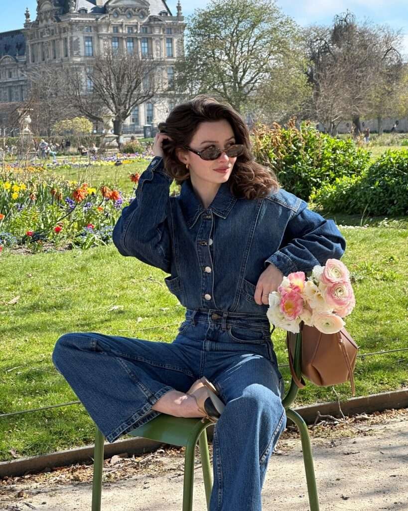 Stylish woman with wavy brown hair wearing a cropped blue denim jacket with buttons, high-waisted wide-leg denim jeans belted at the waist, dark sunglasses, and nude sandals, sitting on a green metal chair in a lush spring garden filled with colorful flowers, holding a beautiful bouquet of pink and white ranunculus, evoking a chic and relaxed spring aesthetic for 2026.