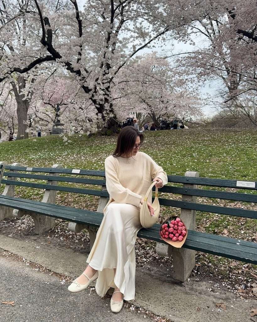 Graceful woman with long dark hair and glasses sitting sideways on a green park bench, wearing an oversized beige cashmere sweater, elegant cream silk maxi skirt with a subtle high-low hem, delicate cream ballet flats, holding a structured beige leather handbag, with a basket of vibrant pink tulips placed beside her, surrounded by lush pink cherry blossom trees in full bloom in a scenic park, evoking a peaceful and romantic neutral spring aesthetic for 2026.
