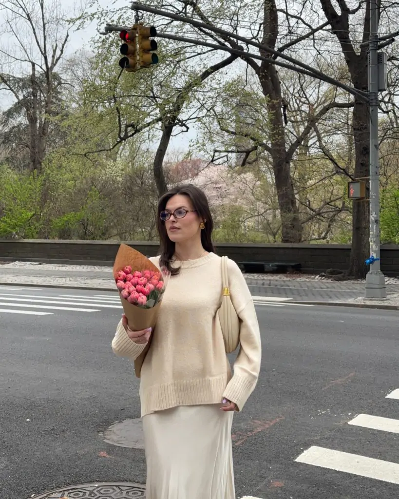 Confident woman with long dark hair wearing oversized cream knit sweater, flowing beige satin maxi skirt, clear-frame glasses, gold hoop earrings, and a structured beige shoulder bag, standing at a city crosswalk holding a large bouquet of vibrant pink tulips wrapped in kraft paper, with blooming cherry blossom trees and urban greenery in the background, embodying a sophisticated neutral spring aesthetic for 2026.