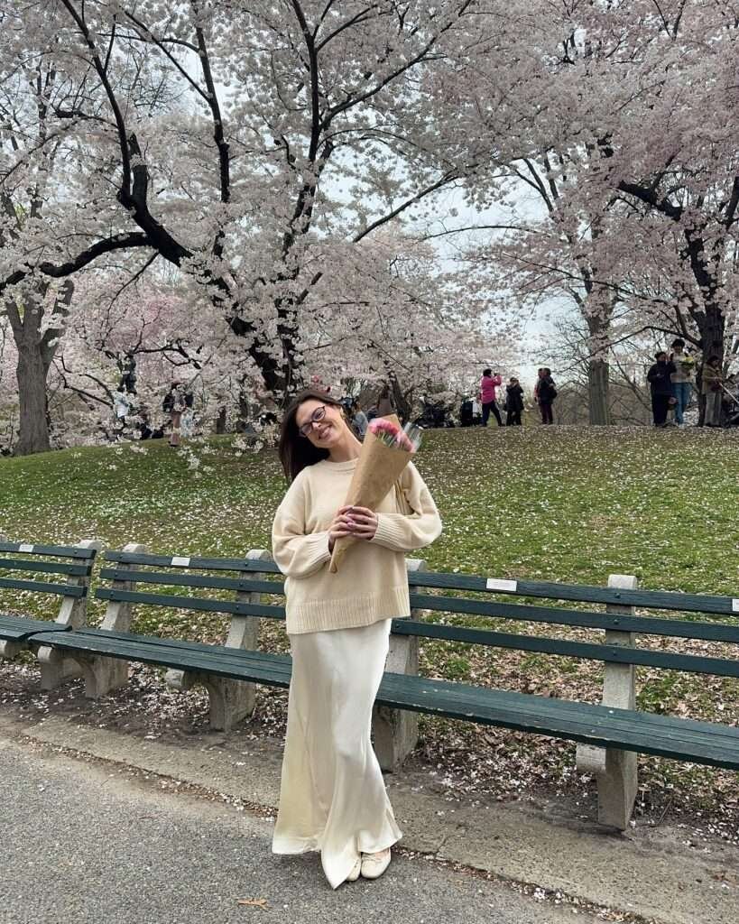 Smiling woman with dark hair and glasses wearing an oversized cream knit sweater, flowing ivory satin maxi skirt, and matching cream ballet flats, standing on a grassy path in a park surrounded by vibrant pink cherry blossom trees in full bloom, holding a bouquet of pink flowers wrapped in kraft paper, capturing a soft and romantic spring aesthetic for 2026.