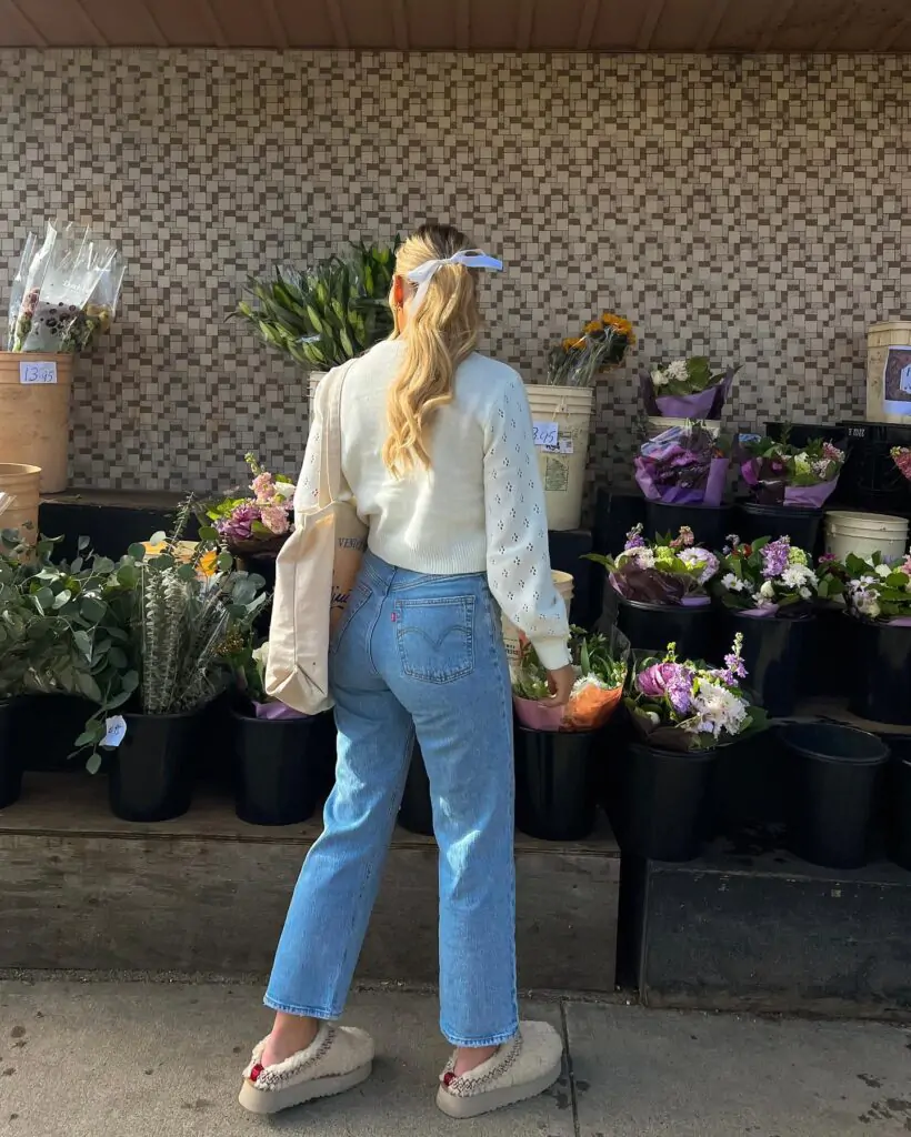 Obsessed with this fresh and fun spring look for 2026: a textured cream eyelet cardigan layered over high-waisted light wash wide-leg jeans, topped off with a blue bow in the hair, a casual tote, and super-cute embellished platform clogs. Back view at the flower market surrounded by vibrant potted blooms and greenery makes it feel so alive and seasonal – the perfect casual spring outfit for market days, casual hangouts, or sunny errands. Pin this effortless spring aesthetic and get ready to copy!