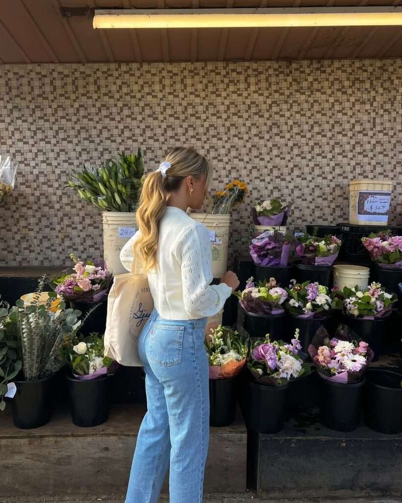This fresh spring moment is made for pinning: a lightweight white eyelet cardigan with delicate details layered over timeless high-waisted blue jeans, finished with a simple ponytail bow and a sturdy canvas tote. Surrounded by vibrant buckets of spring flowers at the market, the outfit perfectly captures that effortless cute spring outfit energy – think weekend errands, coffee runs, or sunny market days. Save this easy, feminine spring 2026 inspiration and add a touch of romance to your wardrobe!