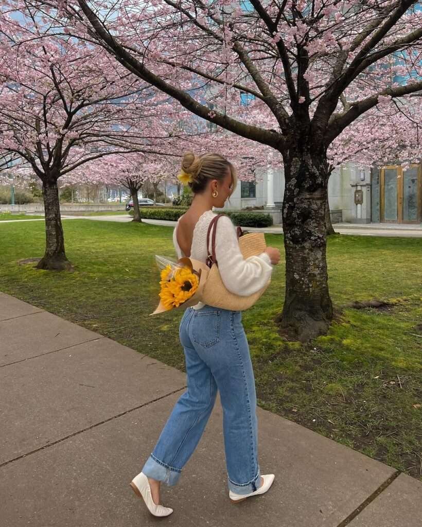 This sunny spring walk is total 2026 inspiration: a breezy white lace sweater with an open back detail flows over high-waisted light wash wide-leg jeans, finished with crisp white ballet flats, a yellow flower in the bun, gold hoops, and a textured straw tote holding cheerful sunflowers. Captured strolling beneath pink cherry blossom branches in full bloom, the outfit radiates pure joy and casual spring fashion elegance – save this romantic, effortless look for your next flower-filled outing or weekend adventure.