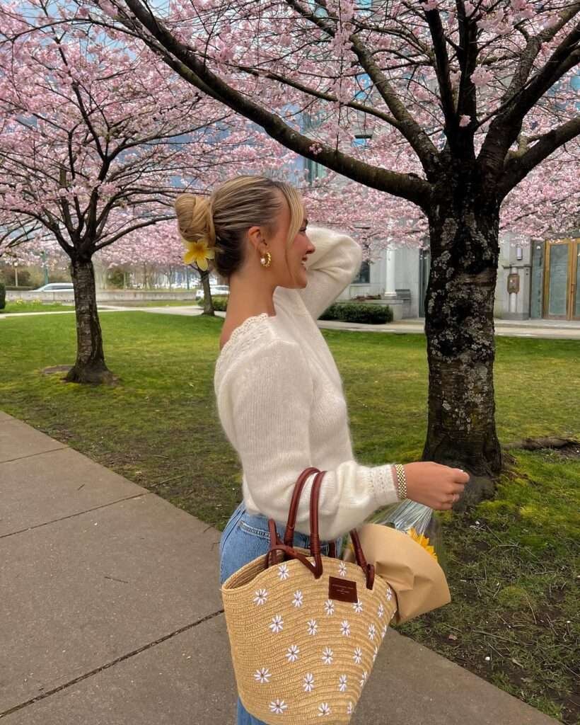 Blonde woman with hair in a high messy bun adorned with a yellow flower, smiling over her shoulder while standing under vibrant pink cherry blossom trees in an urban park, wearing an off-shoulder white open-back lace knit sweater, high-waisted light blue jeans, gold hoop earrings, and holding a large bouquet of bright yellow sunflowers wrapped in kraft paper plus a woven straw tote bag with daisy embroidery and brown leather handles, capturing a joyful and romantic spring aesthetic for 2026.