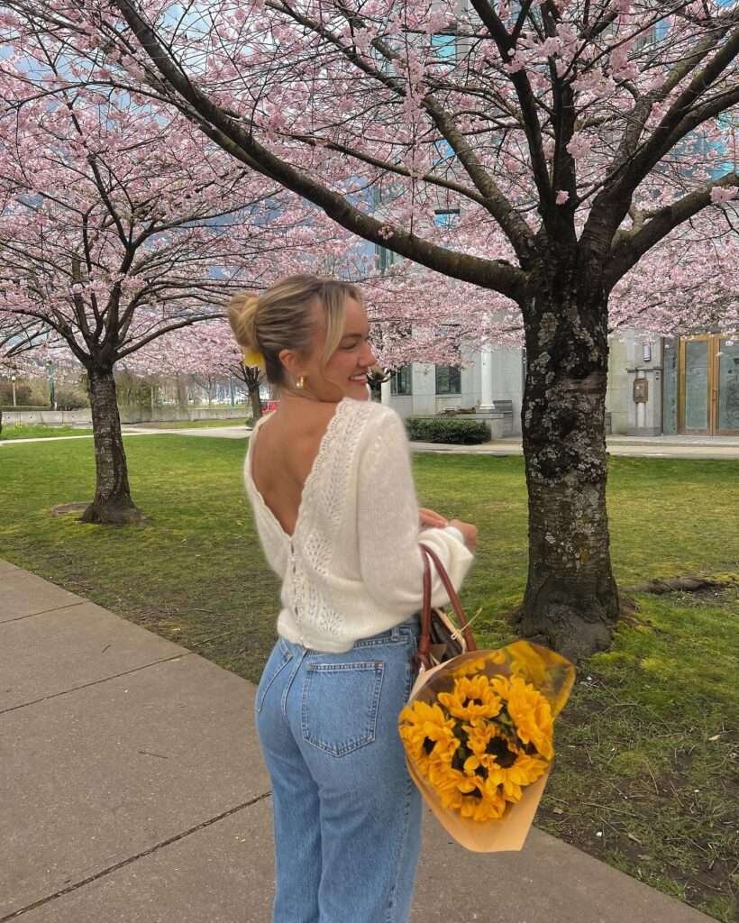 Blonde woman with hair in a high messy bun adorned with a yellow flower, smiling over her shoulder while standing under vibrant pink cherry blossom trees in an urban park, wearing an off-shoulder white open-back lace knit sweater, high-waisted light blue jeans, gold hoop earrings, and holding a large bouquet of bright yellow sunflowers wrapped in kraft paper plus a woven straw tote bag with daisy embroidery and brown leather handles, capturing a joyful and romantic spring aesthetic for 2026.