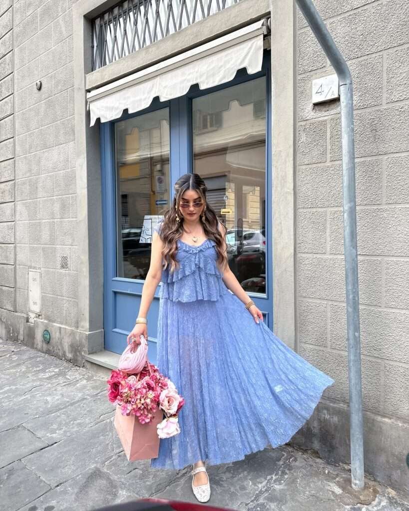 Radiant woman with long wavy dark hair and rose-tinted oval sunglasses standing gracefully on a sunny European street outside a charming blue-shuttered shop, wearing a sleeveless light blue tiered midi dress with delicate ruffles, sheer textured fabric, and a flowy pleated skirt, paired with white pointed-toe ballet flats with subtle sparkle, gold jewelry including hoop earrings and layered necklaces, and holding a large pink shopping bag filled with vibrant pink and peach roses and hydrangeas, embodying a romantic, ethereal, and feminine spring aesthetic for 2026.