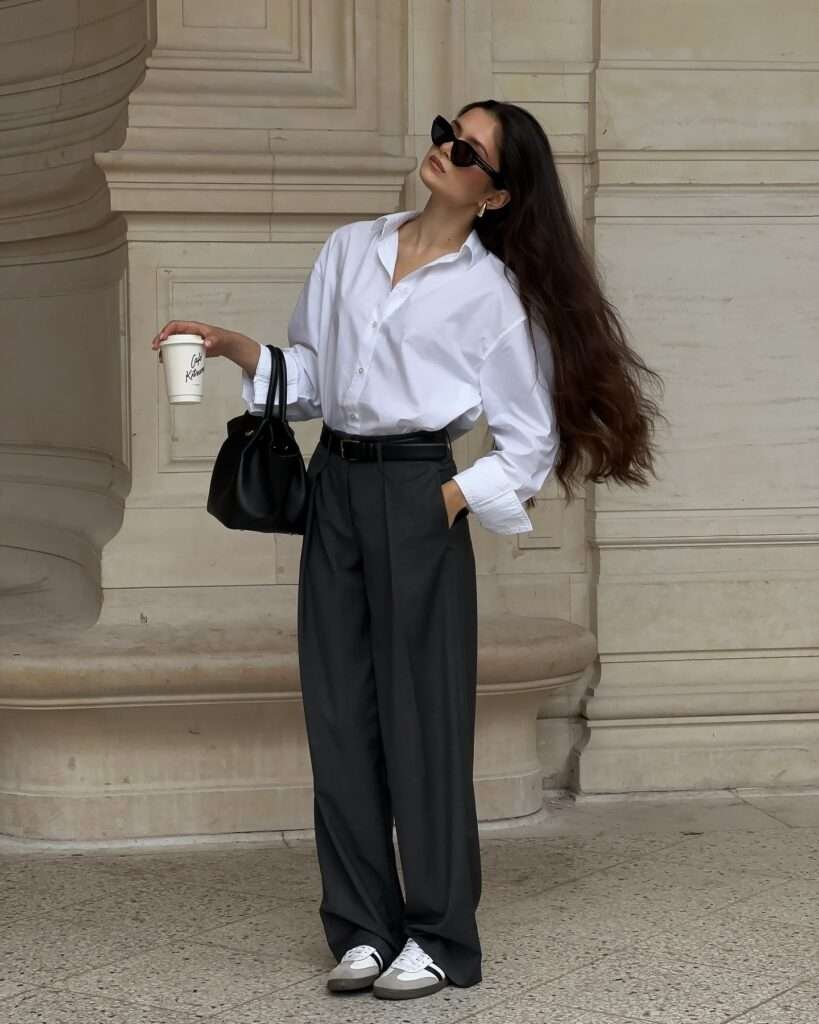 This elevated yet easy spring ensemble is 2026 perfection: a clean white oversized button-down shirt contrasts beautifully with high-waisted charcoal pleated trousers cinched by a black belt, grounded with chunky white sneakers and finished with a sleek black bag and coffee in hand. Captured leaning against classic architecture with a relaxed, confident pose, the look perfectly captures that polished spring aesthetic – ideal for work-to-weekend transitions, city strolls, or effortless sophistication.