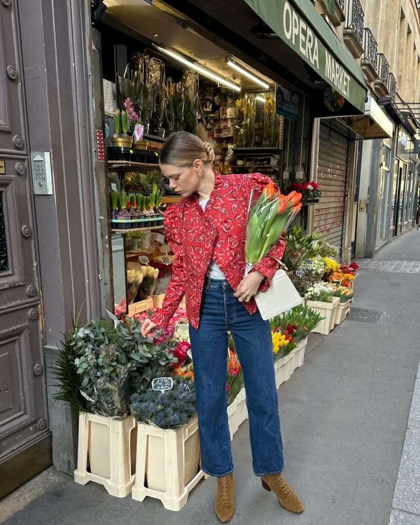 Loving this vibrant spring statement for 2026: a ruffled red paisley jacket brings rich color and texture over a simple white tee and classic wide-leg blue jeans, finished with suede lace-up boots and a sweet bouquet of orange tulips. Posed at a picturesque flower market surrounded by blooming crates and elegant Paris streets, the outfit blends playful pattern with timeless denim for that ultimate spring aesthetic – save it for your next market day, weekend outing, or to add some joyful energy to your wardrobe.