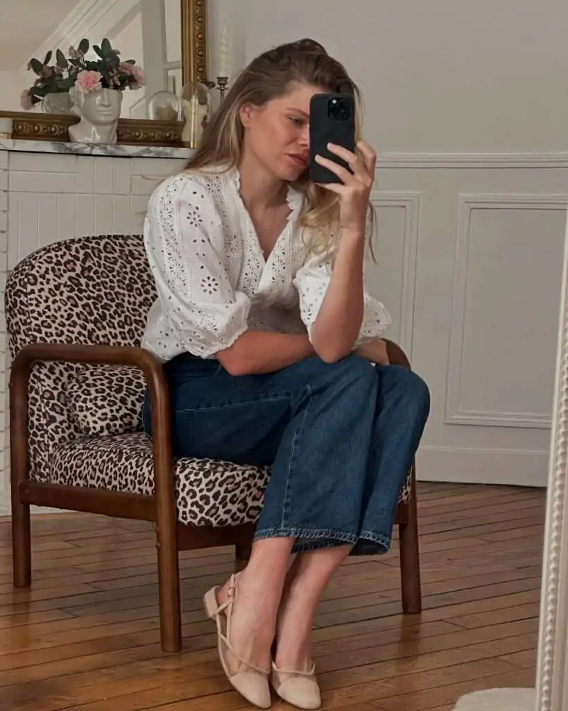 Blonde woman with long wavy hair taking a mirror selfie in a cozy, light-filled room with paneled walls and vintage decor, wearing a breezy white eyelet embroidered blouse with voluminous three-quarter sleeves and a deep V-neck, high-waisted dark wash denim midi skirt, nude strappy ballet flats, seated casually on a leopard-print upholstered wooden chair, capturing a soft, feminine, and relaxed spring aesthetic for 2026.