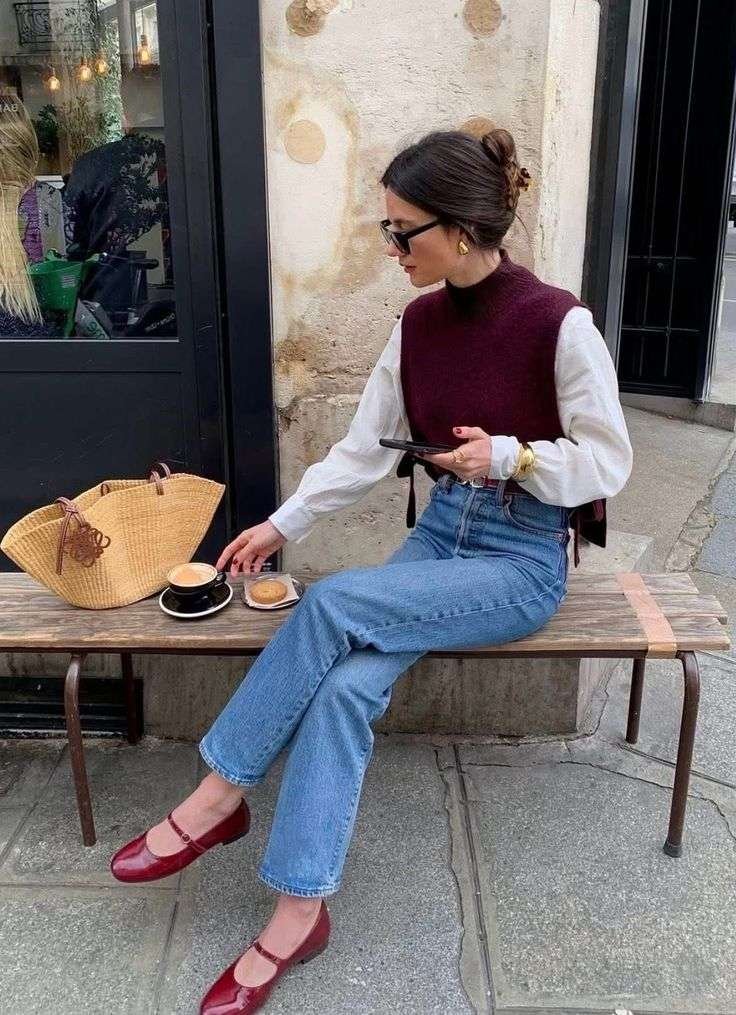 A dark red or burgundy sleeveless knit top, often styled with a mock neck or round neckline. It is layered over a crisp white long-sleeved blouse with button cuffs. High-waisted, straight-leg denim in a classic blue wash. Similar styles include the Brandy Melville Quinn Jeans. Patent leather Mary Jane ballet flats in a deep red or berry shade. The specific pair shown is highly similar to the "Camélia" signature ballerinas from Pied De Biche or the Charles & Keith Carmel patent buckled flats.