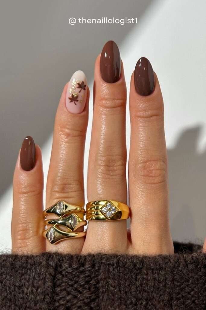 Four fingers in glossy brown with one stunning accent nail — a nude base adorned with a brown maple leaf and gold foil flakes. Peak autumn energy.