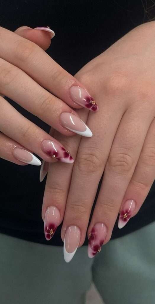 Elevate the timeless French tip by pairing crisp white smiles with a masterful, soft-focused floral watercolor effect on select accent nails. Deep burgundy petals bleed effortlessly into a sheer base, punctuated by tiny gold caviar beads at the center for a subtle touch of jewelry-like hardware. The sleek, elongated almond silhouette provides a striking canvas, making this highly artistic set look like a bespoke masterpiece straight from a luxury salon.