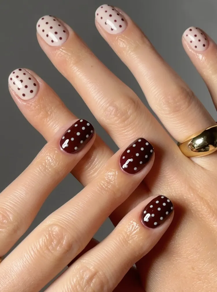 Short round nails with tiny polka dots, alternating between a nude base with burgundy dots and a burgundy base with white dots.