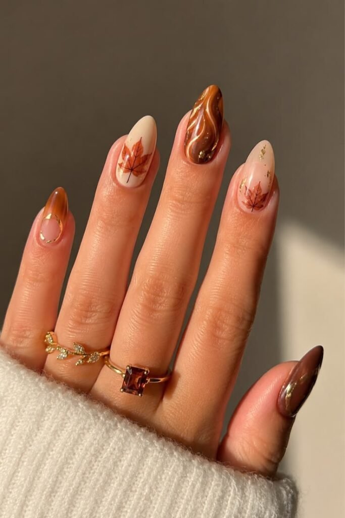 Almond nails in warm nude and brown tones featuring hand-painted maple leaf nail art, a chrome swirl design, and gold foil flakes.