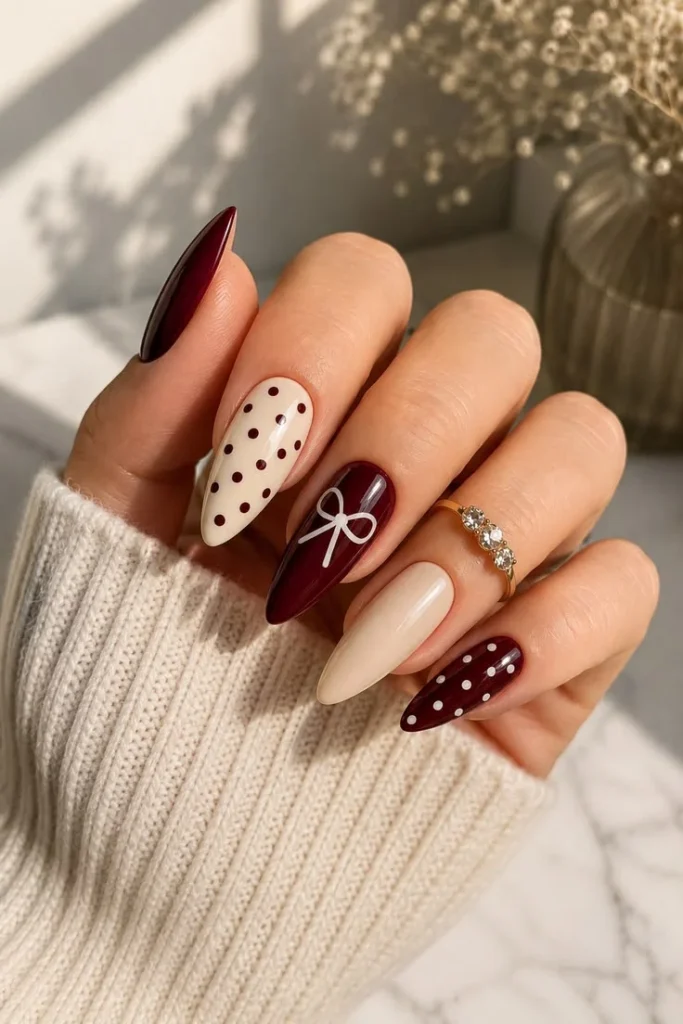 Long almond nails in a mismatched burgundy and cream manicure featuring polka dots and a hand-painted white bow, worn with a cream sweater.
