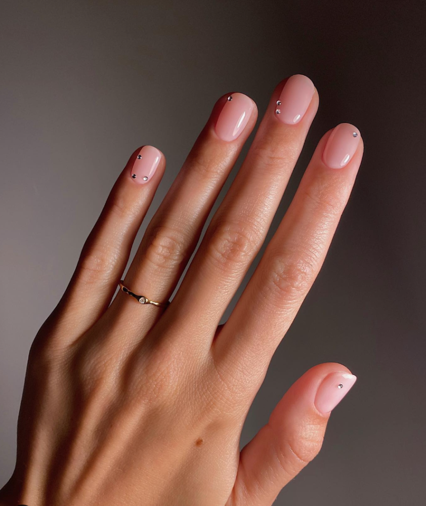 Short oval nails in a glossy nude polish with a few small crystal accents scattered across different nails.