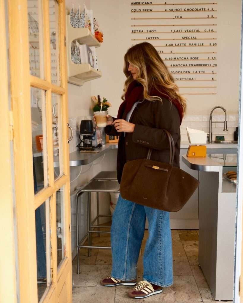 Woman in an oversized brown blazer coat and wide-leg jeans holding an iced coffee inside a cafe