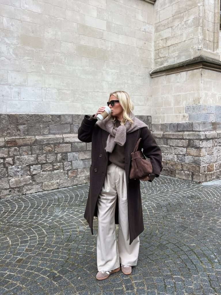 Woman drinking coffee in a brown wool coat and champagne satin wide-leg trousers on a cobblestone street