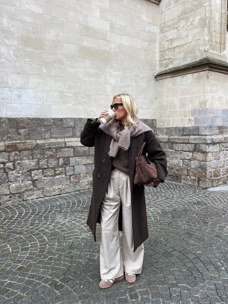 Woman drinking coffee in a brown wool coat and champagne satin wide-leg trousers on a cobblestone street