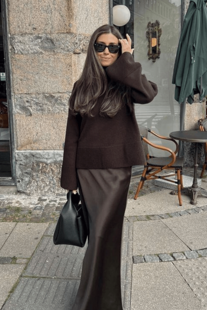 Woman in an oversized brown knit sweater and brown satin maxi skirt standing on a city street