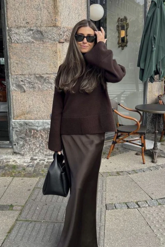 Woman in an oversized brown knit sweater and brown satin maxi skirt standing on a city street