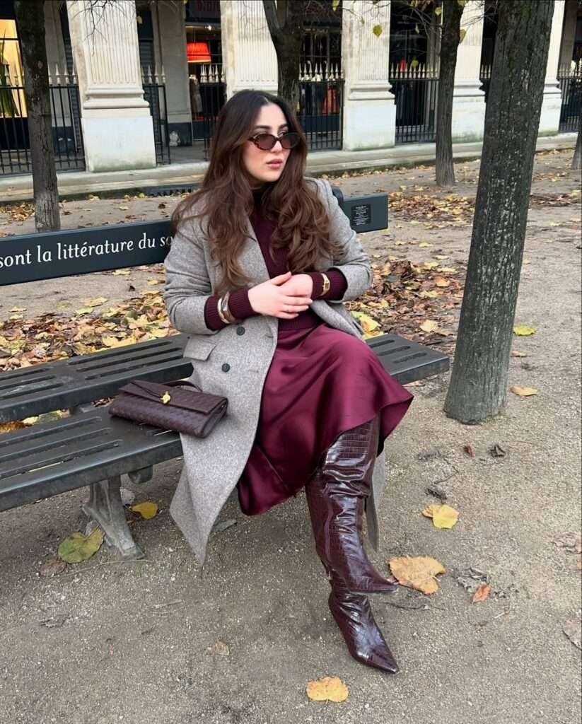 Woman sitting on a park bench wearing a burgundy turtleneck, satin skirt, and knee-high boots under a grey coat