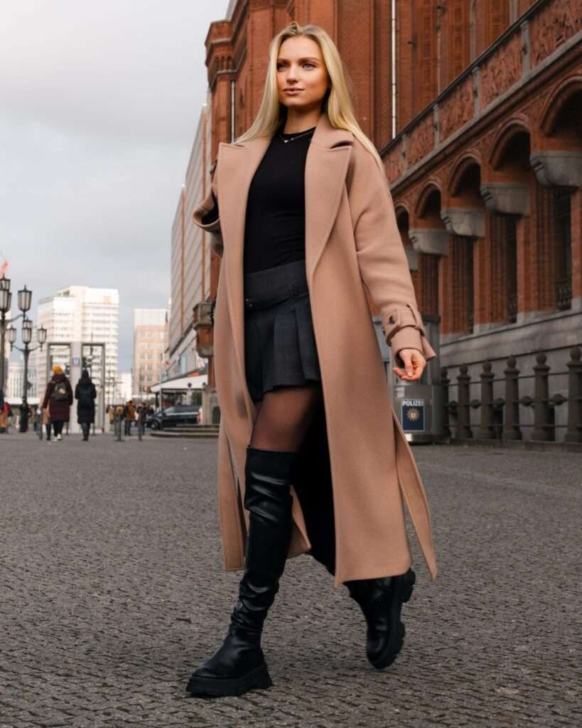 Woman walking in a camel trench coat over a plaid mini skirt and tall black boots