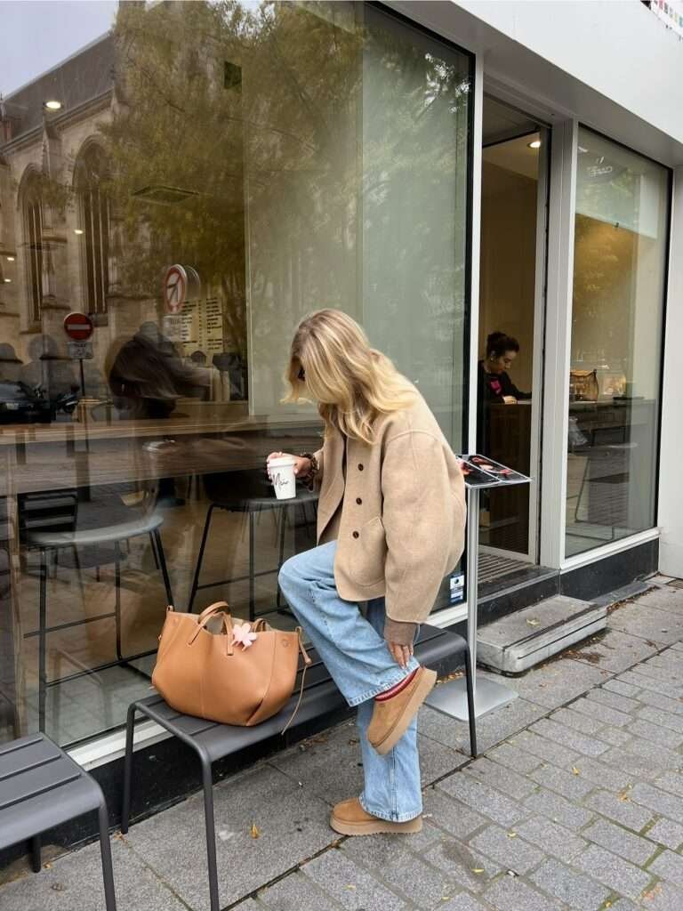 Woman in a camel jacket and wide-leg jeans stepping onto a bench outside a café